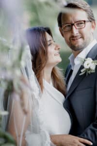 Braut und Bräutigam stehen dicht beieinander, umarmt von weichgezeichnetem Grün. Die Braut in weißem Kleid und Schleier blickt den Bräutigam an, der eine Brille und einen dunklen Anzug mit einer weißen Blumenboutonniere trägt. Beide lächeln sanft an diesem wunderschönen Hochzeitstag.