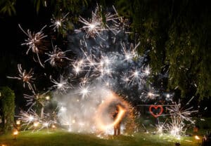 Feuerwerk erhellt den Nachthimmel über einer Grasfläche mit einer leuchtenden Herzform im Hintergrund und ruft die Magie einer Hochzeit hervor. Eine Person steht umgeben von Wunderkerzen und erzeugt ein dynamisches Schauspiel aus Licht und Energie, perfekt für diese besonderen Momente bei der Hochzeitsplanung.