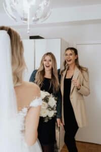 Eine Braut in einem weißen Kleid steht zwei Frauen gegenüber, die in schwarzen Outfits mit Jacke und Blazer gekleidet sind. Eine der Frauen lächelt strahlend und hält einen Blumenstrauß in der Hand, der das elegante Hochzeitsambiente in dem gut beleuchteten, mit einem Kronleuchter geschmückten Raum ergänzt.