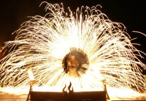 Ein Darsteller, perfekt für eine Hochzeitsfeier, steht in einem rotierenden Apparat aus Stahlwolle und erzeugt ein schillerndes, feuerwerksähnliches Schauspiel aus Funken, die in einem kreisförmigen Muster vor dem dunklen Hintergrund nach außen strahlen. Ideal für alle, die ihren Hochzeitsplänen etwas Magie verleihen möchten.