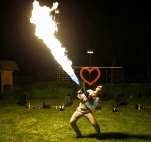 Eine Person in heller Kleidung steht nachts im Freien und schießt mit einem Flammenwerfer eine große Flamme in die Luft. Hinter ihr, auf einer für eine extravagante Hochzeit geschmückten Bühne, steht eine rote herzförmige Dekoration.