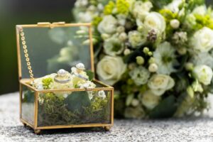 Eine goldene Schmuckschatulle mit Glasdeckel enthält zwei Ringe, eingebettet in Moos und kleine weiße Blumen, die die perfekte Hochzeit symbolisieren. Sie ruht auf einer Steinoberfläche neben einem üppigen Strauß weißer und grüner Blumen und erinnert an die Eleganz einer sorgfältigen Hochzeitsplanung.