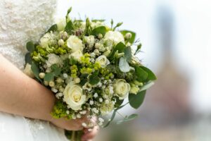 Eine Braut, die einen Strauß weißer Rosen, grüner Blätter und kleiner weißer Blumen hält, zeigt mit ihrem eleganten, natürlichen Look die Schönheit einer Hochzeit. Die Vielfalt an Texturen und sanften Farben des Blumenarrangements verleiht der Hochzeitsszene vor einem verschwommenen Hintergrund eine perfekte Note.