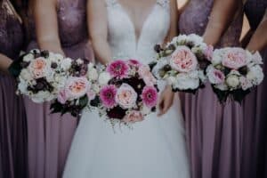 Brautjungfern und Braut halten Sträuße mit rosa und weißen Blumen. Sie tragen malvenfarbene Spitzenkleider, während die Braut in einem weißen Kleid umwerfend aussieht. Der Fokus liegt auf diesen lebendigen Blumenarrangements, perfekt für jede Hochzeitsplanung.