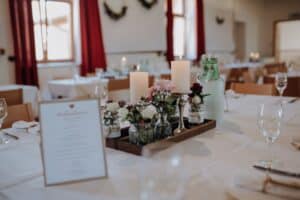 Ein wunderschön gedeckter Esstisch mit weißen Tischdecken, perfekt für eine Hochzeit, mit einem Arrangement aus Kerzen und Blumen in eleganten Vasen. Eine Menükarte liegt auf dem Tisch, neben leeren Weingläsern und einer Wasserflasche. Rote Vorhänge und Kränze schmücken den Hintergrund.