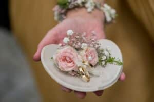 Eine herzförmige Schale hält elegant zwei goldene Ringe, die mit rosa Rosen, Schleierkraut und Grün geschmückt sind und die Essenz der Hochzeit verkörpern. Dieses zarte Arrangement ruht auf einer ausgestreckten Hand und fängt den romantischen Geist der Hochzeit wunderbar ein.