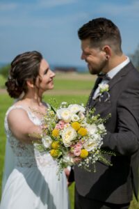 Braut und Bräutigam lächeln sich im Freien an, während sie in ihre Hochzeit vertieft sind. Die Braut hält einen Strauß gelber, weißer und rosa Blumen in der Hand, während der Bräutigam einen dunklen Anzug mit einer Ansteckblume trägt. Hinter ihnen erstreckt sich eine grüne Wiese unter dem strahlend blauen Himmel.