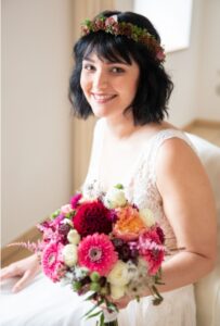 Eine Braut mit dunklem Haar sitzt lächelnd da, trägt eine Blumenkrone und ein Spitzenbrautkleid und genießt die Freude ihrer Hochzeit. Sie hält einen bunten Strauß rosa, roter und weißer Blumen in den Händen. Der Hintergrund ist sanft beleuchtet und verschwommen und fängt die Essenz von Heiraten wunderschön ein.