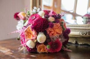 Ein farbenfroher Blumenstrauß, perfekt für eine Hochzeit, besteht aus einer Mischung aus rosa, orangefarbenen und weißen Blüten mit grünen Akzenten und steht auf einem Holztisch vor einem verzierten Spiegel. Das üppige Arrangement reflektiert das Licht wunderschön auf der Oberfläche des Spiegels.