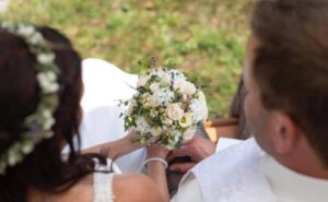 Braut und Bräutigam sitzen eng beieinander, halten sich an den Händen und sind in ihre Hochzeit vertieft. Der Brautstrauß voller weißer und pastellfarbener Blumen ist im Fokus. Sie sind draußen, umgeben von leuchtendem Grün. Die Braut trägt einen Blumenkopfschmuck, der die Essenz ihres besonderen Tages einfängt.