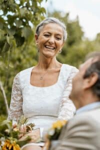 Eine ältere Frau in einem weißen Spitzenkleid lächelt eine im Freien sitzende Person herzlich an und verkörpert damit die Freude am Heiraten. Sie hält einen Blumenstrauß in der Hand, umgeben von grünem Laub unter einem hellen Himmel, als würde sie die Essenz einer sorgfältigen Hochzeitsplanung verkörpern.