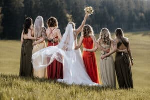 Eine Braut in einem weißen Kleid mit langem Schleier steht auf einem Feld, umgeben von sechs Brautjungfern in bunten Kleidern. Sie stehen mit dem Rücken zur Kamera, wobei die Braut einen Blumenstrauß hochhält. Dieser perfekte Moment von Heiraten fängt die Schönheit der Natur mit einem Wald im Hintergrund ein.