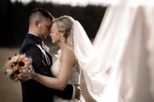 Braut und Bräutigam umarmen sich im Freien und legen sanft ihre Stirnen aneinander. Dies verkörpert die Essenz einer perfekten Hochzeit. Die Braut hält einen Strauß Sonnenblumen und ihr Schleier weht elegant im Wind vor einer verschwommenen Naturkulisse.
