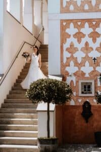 Eine Braut in einem weißen Brautkleid geht mit einem Blumenstrauß in der Hand eine Treppe hinunter und verkörpert damit den Geist einer Hochzeit. Die Kulisse besticht durch kunstvolle architektonische Details und eine dekorative Wand mit geometrischen Mustern. Topfpflanzen säumen den Weg und unterstreichen diese wunderschöne Hochzeitsszene.