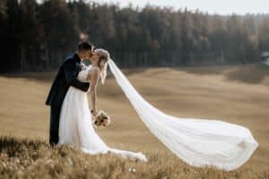 Braut und Bräutigam küssen sich auf einer sonnenbeschienenen Wiese, ihre Hochzeit wird von der Schönheit der Natur eingerahmt. Die Braut hält einen Blumenstrauß, während ihr langer Schleier im Wind weht. In der Ferne runden Bäume diesen bildschönen Moment für eine unvergessliche Hochzeitsplanung ab.