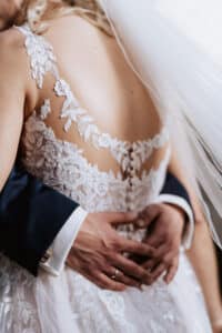Eine Nahaufnahme von Braut und Bräutigam, die sich während ihrer Hochzeit umarmen. Das Spitzenbrautkleid der Braut weist aufwendige Blumendetails und Knöpfe am Rücken auf. Die Hand des Bräutigams ruht sanft auf ihrer Taille und zeigt seinen Ehering.