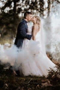 Inmitten einer Waldkulisse, eingehüllt in Nebel und sanftes Sonnenlicht, küssen sich Braut und Bräutigam romantisch. Das weiße Kleid und der Schleier der Braut bilden einen schönen Kontrast zum dunklen Anzug des Bräutigams. Diese ätherische Szene fängt die Essenz einer unvergesslichen Hochzeit ein.
