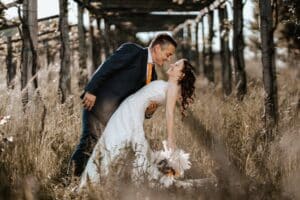 Ein Paar in Hochzeitskleidung teilt einen freudigen Moment in einer rustikalen Umgebung im Freien. Der Bräutigam beugt sich über die Braut, die einen Blumenstrauß hält, unter einer hölzernen Pergola, die von hohem Gras umgeben ist – eine perfekte Szene für Hochzeitsträume voller Sonnenlicht und Grün.