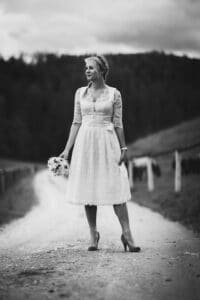 Eine Frau in einem knielangen Kleid und High Heels steht mit einem Blumenstrauß in der Hand auf einer Landstraße und fängt damit die Essenz von „Hochzeit“ ein. Die Außenkulisse mit Bäumen und Hügeln im schwarz-weißen Hintergrund verleiht diesem Moment der Liebe und „Heiraten“ eine zeitlose Note.