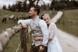 Ein Paar in Hochzeitskleidung steht umarmt neben einem rustikalen Zaun und verkörpert die Romantik ihrer Hochzeit. Die Frau legt ihren Kopf mit zufriedenem Gesichtsausdruck auf den Rücken des Mannes, während Pferde in der Nähe grasen und ein Feldweg in die idyllische Ferne führt.