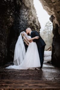 Braut und Bräutigam küssen sich in einer malerischen Umgebung im Freien, perfekt für alle, die von ihrer Hochzeit träumen. Die Braut ist in einem weißen Kleid und Schleier atemberaubend, während der Bräutigam in seinem dunklen Anzug auf einem Holzsteg, der von majestätischen Felsformationen und Bäumen gesäumt ist, schick aussieht.