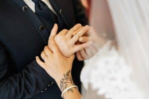 Nahaufnahme einer Braut, die dem Bräutigam während der Hochzeit einen Ehering an den Finger steckt. Der Bräutigam trägt einen schwarzen Anzug und auf der Hand der Braut ist ein Blumentattoo zu sehen. Der Fokus liegt auf den Händen, die den Ringtausch bei dieser wunderschönen Zeremonie symbolisieren.