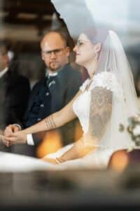 Während der Hochzeitszeremonie enthüllt eine Braut in einem weißen Kleid mit Spitzendetails und einem Schleier ihren tätowierten Arm. Dicht neben ihr sitzt der Bräutigam im dunklen Anzug und blickt nach vorne. Dieser heitere Moment verkörpert die Essenz von Liebe und sorgfältiger Hochzeitsplanung.