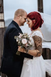 Ein Paar steht Stirn an Stirn und lächelt. Die Braut hält einen Blumenstrauß in der Hand, trägt ein Spitzenkleid mit sichtbaren Tattoos und rotem Haar. Der Bräutigam mit Brille und dunklem Anzug ergänzt sie perfekt. Eine Holzkonstruktion und ein bewölkter Himmel evozieren den romantischen Charme ihrer Hochzeitsfeier.