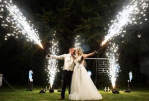 Braut und Bräutigam küssen sich während ihrer Hochzeit mit Wunderkerzen in der Hand. Sie stehen nachts im Freien und hinter ihnen ist ein großes Feuerwerk zu sehen. Die Braut trägt ein weißes Kleid und der Bräutigam einen hellen Anzug. Die festliche Szene wird durch das leuchtende Feuerwerk darüber hell erleuchtet.