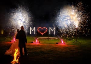 Ein Paar steht nachts draußen und schaut sich das Feuerwerk über einer Lichtinstallation an, auf der hell erleuchtet „M ♥ M“ steht. Dies schafft eine bezaubernde Atmosphäre, die perfekt für eine Hochzeit ist. Eine kleine Feuerstelle sorgt im Vordergrund für Wärme, während hinter den Lichtern ein Springbrunnen leuchtet.