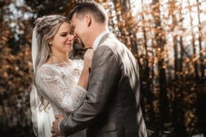 In einem sonnenbeschienenen Wald umarmt sich ein Paar in Hochzeitskleidung und verkörpert den Traum ihrer sorgfältig ausgearbeiteten Hochzeitsplanung. Die Braut in einem Spitzenkleid und einem Haarschmuck mit Blumenmuster blickt den Bräutigam im grauen Anzug inmitten goldener Herbstblätter liebevoll an.