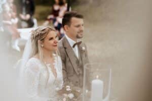 Braut und Bräutigam sitzen während ihrer Hochzeit Seite an Seite. Die Braut trägt ein Spitzenkleid mit Schleier und hält einen Blumenstrauß, während der Bräutigam einen grauen Anzug mit Fliege trägt. Vor ihnen steht eine weiße Kerze, die die Gelübde symbolisiert, die sie an diesem wunderschön geplanten Tag der Hochzeit ausgetauscht haben.