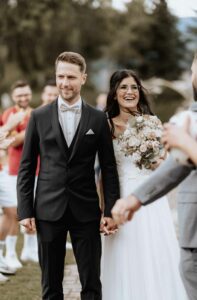 Braut und Bräutigam lächeln, während sie Hand in Hand draußen gehen und ihre Hochzeit feiern. Der Bräutigam trägt einen schwarzen Anzug und die Braut ein weißes Kleid und hält einen Blumenstrauß in der Hand. Im Hintergrund sind Menschen in Freizeitkleidung zu sehen, die freudig klatschen und den freudigen Anlass genießen.