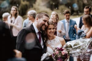 Eine lächelnde Braut und ein Bräutigam umarmen sich bei einer Hochzeit im Freien. Die Braut hält einen Strauß bunter Blumen in der Hand und strahlt vor Freude, während Seifenblasen in der Luft schweben. Gäste in festlicher Kleidung tragen zur festlichen Atmosphäre bei und fangen einen wunderschön geplanten Tag der Liebe und Verbundenheit ein.