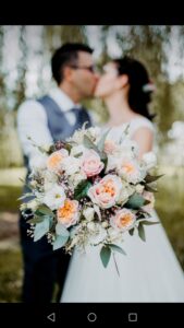 Ein Paar im Hochzeitskleid küsst sich, im Hintergrund leicht verschwommen. Im Fokus steht ein Brautstrauß mit rosa und weißen Rosen und Grünpflanzen, der elegante Hochzeitsplanung verkörpert. Der Hintergrund ist mit weichem, unscharfem Grün bedeckt.