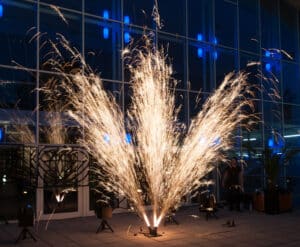 Während einer Hochzeitsfeier schießt ein farbenprächtiges Feuerwerk vor einem modernen Glasgebäude empor und wirft einen warmen Schein auf die reflektierende Oberfläche. Blaue Lichter im Inneren des Gebäudes ergänzen diesen magischen Hochzeitsmoment mit kühlen, kontrastierenden Farbtönen.
