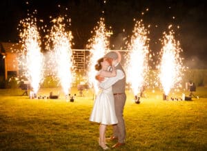 Ein Paar umarmt und küsst sich nachts auf einer Wiese und feiert seine Hochzeit inmitten von hellem Feuerwerk, das in die Luft schießt. Die Frau trägt ein weißes Kleid, während der Mann eine Weste und einen Hut trägt. Ein Hintergrund aus beleuchteten Strukturen verleiht der bezaubernden Szene Magie.