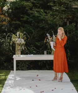Eine Frau in einem orangefarbenen Kleid steht neben einem Tisch auf einer weißen Unterlage im Freien, umgeben von Grün, und ist in die Hochzeitsplanung vertieft. Sie hält Papiere in der Hand und wirkt nachdenklich. Auf dem Tisch stehen große Blumenarrangements und ein Glas Wasser, während Rosenblätter elegant auf der Unterlage verstreut sind.