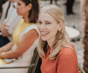 Eine Frau mit langen blonden Haaren, einem korallenfarbenen Oberteil und großen runden Ohrringen sitzt draußen und lächelt in die Kamera, vielleicht in Gedanken versunken über ihre bevorstehende Hochzeit. Im Hintergrund ist eine Person teilweise zu sehen, die ein gelb-weißes Kleid trägt.