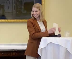 Eine Frau mit blondem Haar, einem braunen Blazer und einem weißen Hemd, steht neben einem Tisch mit weißer Tischdecke. Sie lächelt und hält Papiere in der Hand. Sie strahlt selbstbewusste Expertise in Hochzeitsplanung aus. Auf dem Tisch stehen ein Mikrofon und ein Glas, und ein gerahmtes Gemälde schmückt die Wand hinter ihr.