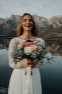 Ein verschwommenes Bild zeigt eine Frau in einem weißen Kleid mit einem Blumenstrauß in der Hand, die vor einem ruhigen See mit majestätischen Bergen im Hintergrund steht – eine perfekte Szene für alle, die davon träumen, inmitten der Schönheit der Natur zu heiraten.