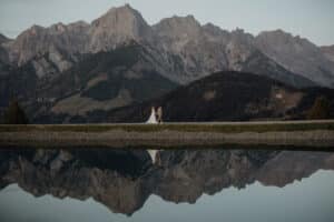 Ein Paar geht an einem ruhigen See entlang, im Wasser ist ihr Spiegelbild zu sehen. Majestätische, schroffe Berge bilden die Kulisse unter einem klaren Himmel und bieten die perfekte Kulisse für ihre Hochzeit. Der Mann trägt einen Anzug, während die Frau ein fließendes weißes Kleid trägt.