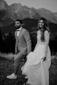 Ein Schwarzweißfoto eines Paares, das seine Hochzeit in einer Berglandschaft feiert. Der Mann trägt einen Anzug und die Frau ein langes, fließendes Kleid. Beide lächeln und wirken glücklich. Die verschwommenen Berge und Bäume tragen zur romantischen Kulisse ihres besonderen Tages bei.