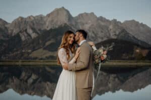 Ein Paar umarmt sich vor einem ruhigen See mit Bergen im Hintergrund und fängt einen perfekten Moment des Hochzeitsglücks ein. Die Braut trägt ein weißes Kleid und hält einen Blumenstrauß, während der Bräutigam einen braunen Anzug trägt. Die Landschaft spiegelt sich im Wasser und schafft eine friedliche Szene.