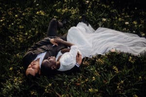 Ein Paar liegt im Gras, umgeben von kleinen gelben Blumen, und verkörpert einen ruhigen und intimen Moment ihrer Hochzeit. In formeller Kleidung gekleidet, die Frau in einem weißen Kleid und der Mann in einem dunklen Anzug, fängt diese Aufnahme von oben die Essenz der Liebe und Feierlichkeit an ihrem besonderen Tag ein.