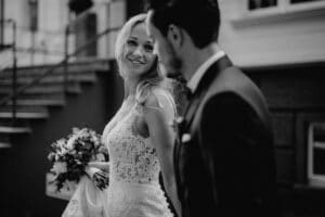 Ein Schwarzweißfoto eines Paares an seinem Hochzeitstag fängt die Essenz des Heiratens ein. Die Braut in einem Spitzenkleid lächelt und hält einen Blumenstrauß in der Hand. Der Bräutigam im Anzug blickt sie an, während sie zusammen an einem Gebäude mit Stufen im Hintergrund vorbeigehen.