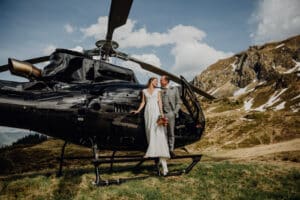 Ein Paar steht auf der Stufe eines schwarzen Hubschraubers, umarmt sich und lächelt. Die Braut in einem weißen Kleid mit einem Blumenstrauß in der Hand und der Bräutigam in einem grauen Anzug feiern ihre Hochzeit inmitten einer Berglandschaft mit Schneeflecken unter einem teilweise bewölkten Himmel.