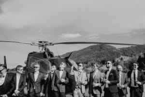Eine Gruppe von Männern in Anzügen steht mit Getränken und Zigarren vor einem Hubschrauber und feiert wahrscheinlich eine extravagante Hochzeit. Die fröhliche Szene spielt sich an einem sonnigen Tag im Freien ab, mit bewaldeten Hügeln im Hintergrund. Das Bild ist in Schwarzweiß gehalten.