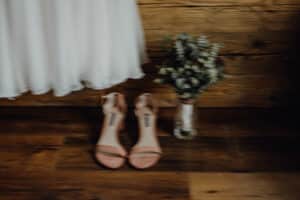 Ein weichgezeichnetes Bild eines weißen Kleids, das anmutig über hautfarbenen High Heels hängt, perfekt für eine Hochzeit. Ein Blumenstrauß in einer Vase verleiht neben den Schuhen Charme. Der rustikale Holzboden und die Holzwand bilden eine ruhige Kulisse für diesen Moment der Hochzeitsplanung.