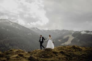 Braut und Bräutigam halten sich an den Händen, während sie über einen felsigen Hügel gehen und verkörpern damit die Essenz einer Hochzeit. In einem weißen Brautkleid und einem schwarzen Anzug gekleidet, strahlt ihre Liebe vor einer dramatischen Kulisse aus Bergen und bewölktem Himmel.